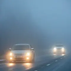 Deux voitures circulent sur une route brumeuse, leurs feux de brouillard avant allumés pour percer le brouillard épais.