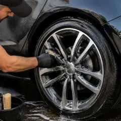 Un homme nettoie des jantes alu avec un produit maison et une brosse. Le pneu est mouillé et savonneux.