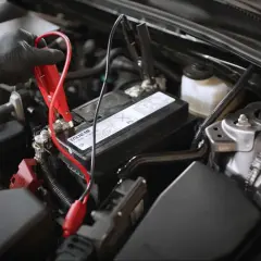 Main d'une personne portant un gant noir connectant des câbles de démarrage à une batterie de voiture, signe d'une batterie qui se décharge.