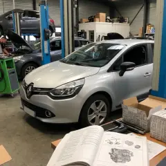 Dans un garage, un mécanicien travaille sur la distribution d'une Clio 4 argentée. Une autre voiture est levée sur un pont.