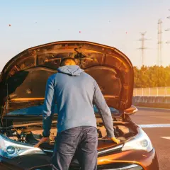 Un homme regarde le moteur de sa voiture, capot ouvert, peut-être un problème de tension batterie voiture. Le soleil brille.