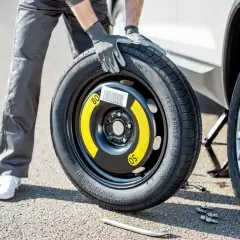 Un homme en gants gris tient une roue de secours pour changer un pneu. La voiture est sur un cric.