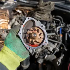 Main d'un mécanicien tenant une pompe à eau de voiture démontée, révélant la turbine rouillée.