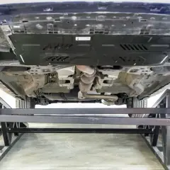 Vue du dessous d'une voiture sur des chandelles. Le système d'échappement est visible, ainsi qu'une plaque de protection. On dirait qu'il y a eu un choc sous la voiture.