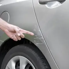 Doigt pointant des éraflures sur une voiture, résultat d'un frottement dans un parking.