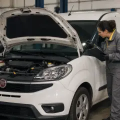Mécanicienne vérifiant le moteur d'un Fiat Doblo blanc. Le capot est ouvert, révélant le compartiment moteur. Elle tient une tablette et un stylo.
