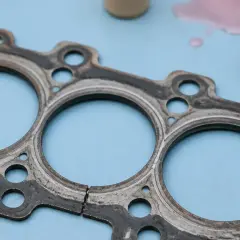 Un joint de culasse usé, prêt à être remplacé. Des traces de liquide de refroidissement rose et un tuyau sont visibles à proximité.