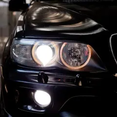 Gros plan sur l'avant d'une voiture noire, les feux de position allumés, créant une lumière intense et des reflets sur la carrosserie.