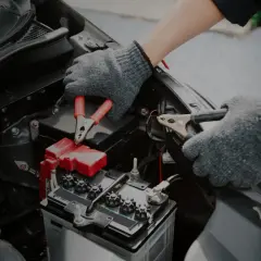 Mains de personne avec des gants gris connectant des câbles à une batterie de voiture. Un symptôme de batterie HS est souvent un démarrage difficile.