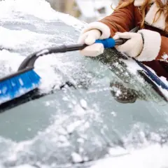 Une personne en manteau brun enlève la neige du pare-brise de sa voiture avec une brosse bleue. Faut il enlever la neige sur la voiture ? Oui, pour la sécurité !
