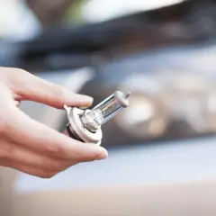 Main tenant une ampoule de phare, se demandant quelle ampoule pour ma voiture. Le capot et le phare de la voiture sont flous en arrière-plan.