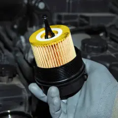 Main d'une personne portant un gant gris tenant un nouveau filtre à huile jaune et un couvercle noir, prêt à le remplacer.