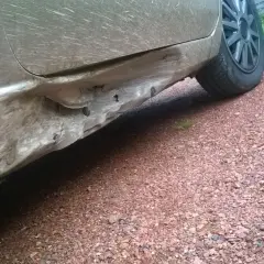 Vue rapprochée d'un bas de caisse enfoncé sur une voiture sale, montrant les dégâts et les fixations exposées.