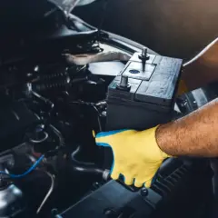 Mains pour gants jaunes tenant une batterie de voiture pour la débrancher. L'intérieur du compartiment moteur est visible.