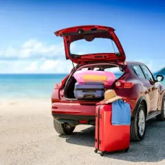 Voiture rouge prête pour le départ en vacances, coffre ouvert rempli de bagages, valise rouge et chapeau de paille sur le sable, mer bleue en arrière-plan.