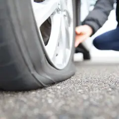 Gros plan sur un pneu crevé, une personne accroupie vérifie les dégâts. La route est asphaltée.