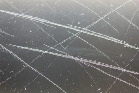 Gros plan sur une vitre très rayée, montrant des lignes fines et des éclaboussures d'eau. Idéal pour illustrer comment réparer une vitre rayée.