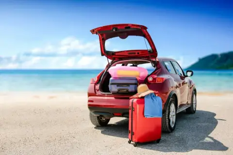 Voiture rouge prête pour le départ en vacances, coffre ouvert rempli de bagages, valise rouge et chapeau de paille sur le sable, mer bleue en arrière-plan.