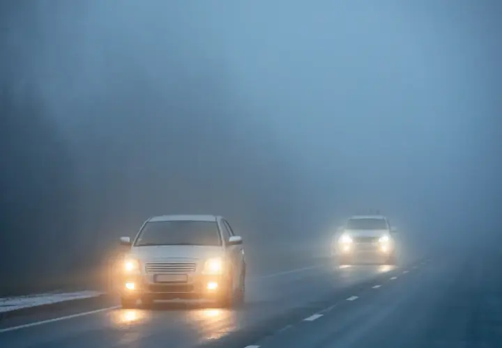 Deux voitures circulent sur une route brumeuse, leurs feux de brouillard avant allumés pour percer le brouillard épais.