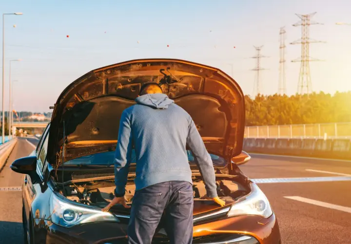 Un homme regarde le moteur de sa voiture, capot ouvert, peut-être un problème de tension batterie voiture. Le soleil brille.
