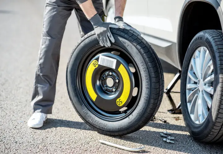 Un homme en gants gris tient une roue de secours pour changer un pneu. La voiture est sur un cric.