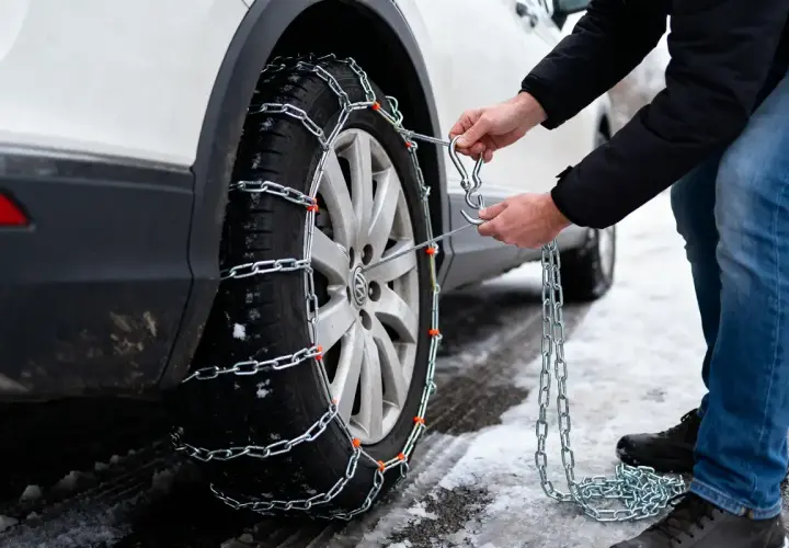 Comment mettre des chaînes neige : un homme attache une chaîne à un pneu de voiture sur une route enneigée.