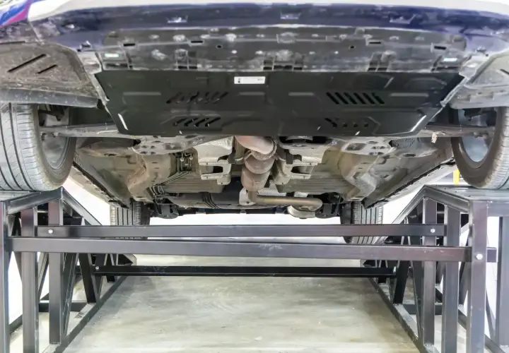 Vue du dessous d'une voiture sur des chandelles. Le système d'échappement est visible, ainsi qu'une plaque de protection. On dirait qu'il y a eu un choc sous la voiture.