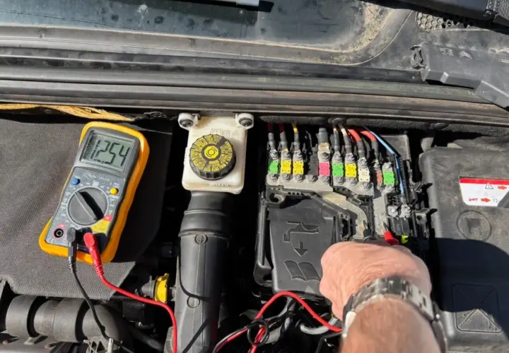 Diagnostic d'une C4 phase 2 : un multimètre affiche 12.54V près du boîtier de fusibles, indiquant une vérification avant de changer la batterie.