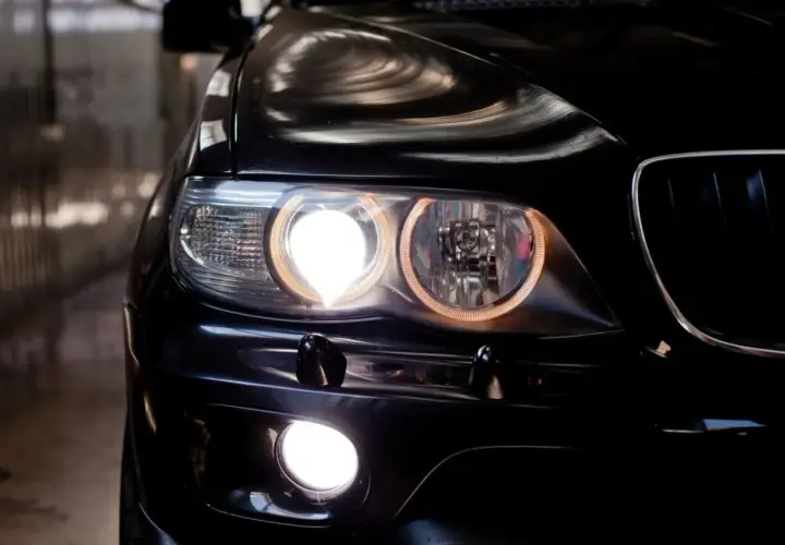 Gros plan sur l'avant d'une voiture noire, les feux de position allumés, créant une lumière intense et des reflets sur la carrosserie.