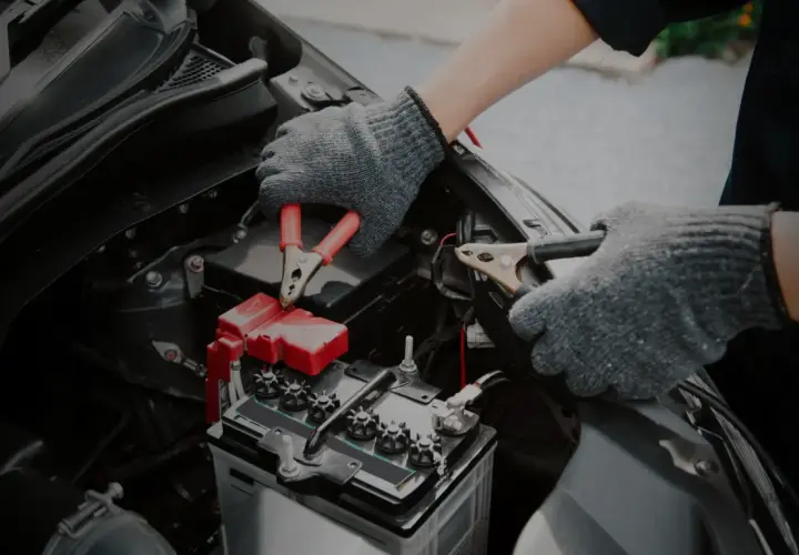 Mains de personne avec des gants gris connectant des câbles à une batterie de voiture. Un symptôme de batterie HS est souvent un démarrage difficile.