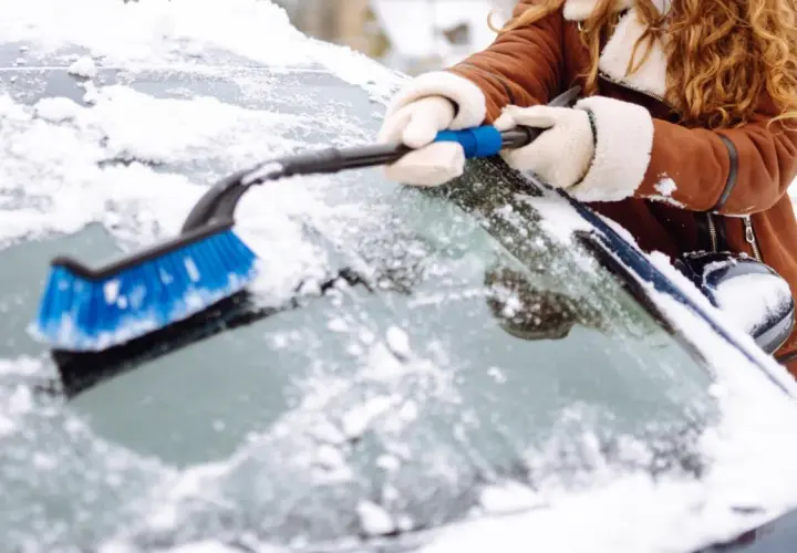 Une personne en manteau brun enlève la neige du pare-brise de sa voiture avec une brosse bleue. Faut il enlever la neige sur la voiture ? Oui, pour la sécurité !
