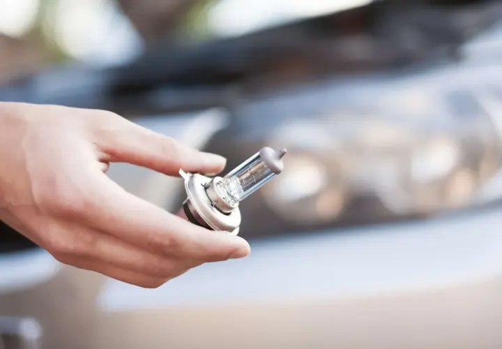 Main tenant une ampoule de phare, se demandant quelle ampoule pour ma voiture. Le capot et le phare de la voiture sont flous en arrière-plan.
