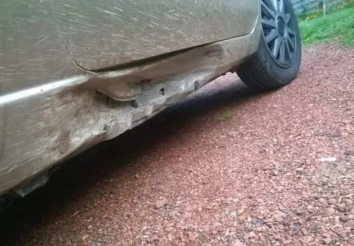 Vue rapprochée d'un bas de caisse enfoncé sur une voiture sale, montrant les dégâts et les fixations exposées.