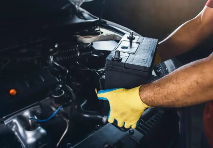 Mains pour gants jaunes tenant une batterie de voiture pour la débrancher. L'intérieur du compartiment moteur est visible.