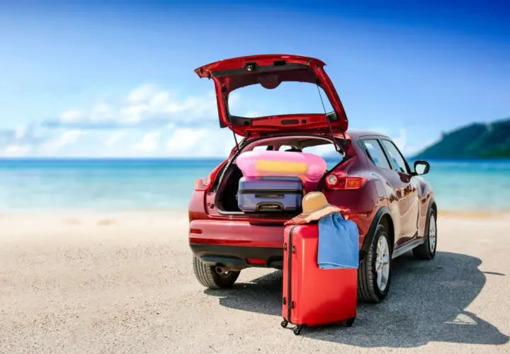 Voiture rouge prête pour le départ en vacances, coffre ouvert rempli de bagages, valise rouge et chapeau de paille sur le sable, mer bleue en arrière-plan.
