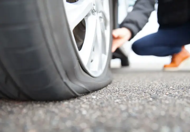 Gros plan sur un pneu crevé, une personne accroupie vérifie les dégâts. La route est asphaltée.