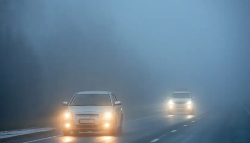 Deux voitures circulent sur une route brumeuse, leurs feux de brouillard avant allumés pour percer le brouillard épais.