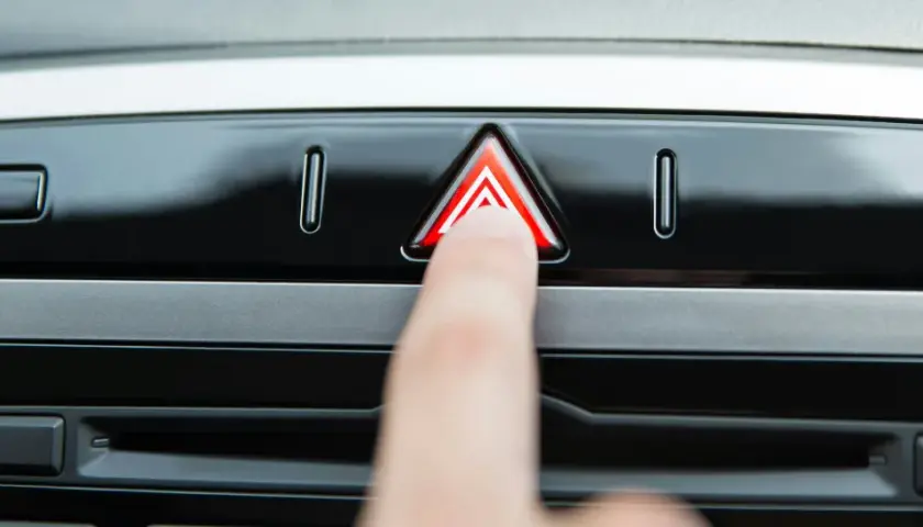 Un doigt appuie sur le bouton des feux de détresse, un triangle rouge lumineux sur le tableau de bord d'une voiture.