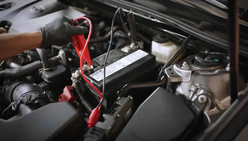 Main d'une personne portant un gant noir connectant des câbles de démarrage à une batterie de voiture, signe d'une batterie qui se décharge.