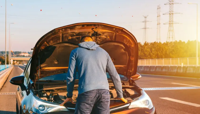 Un homme regarde le moteur de sa voiture, capot ouvert, peut-être un problème de tension batterie voiture. Le soleil brille.