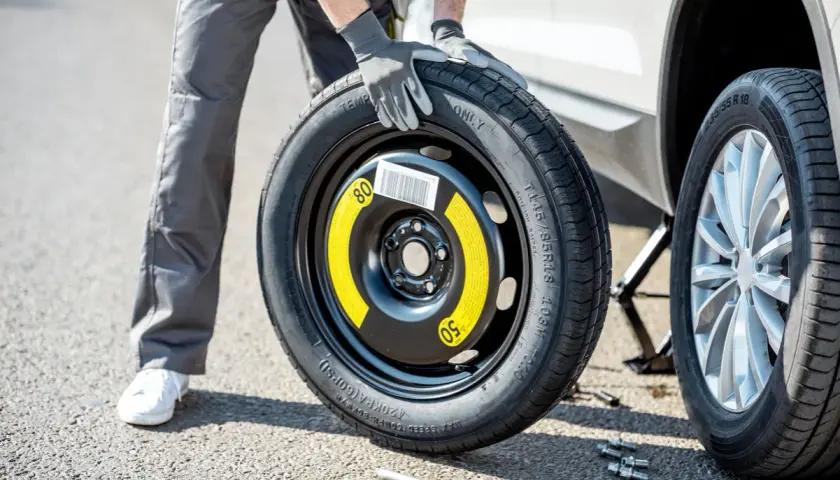 Un homme en gants gris tient une roue de secours pour changer un pneu. La voiture est sur un cric.