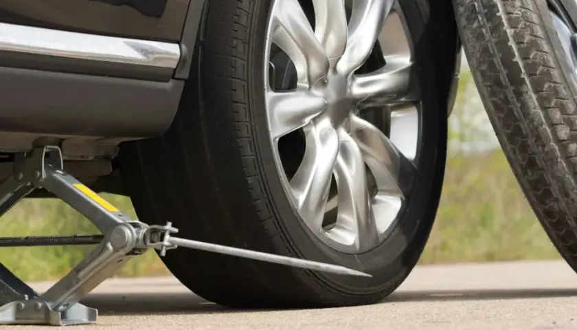 Cric de voiture soulevant un véhicule, avec une roue de secours à proximité.