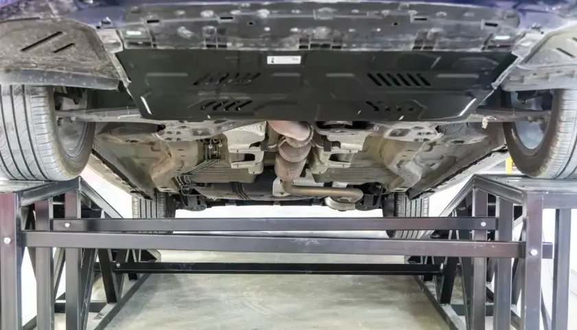 Vue du dessous d'une voiture sur des chandelles. Le système d'échappement est visible, ainsi qu'une plaque de protection. On dirait qu'il y a eu un choc sous la voiture.