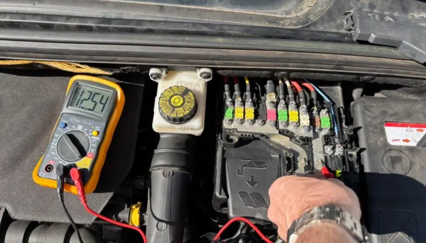 Diagnostic d'une C4 phase 2 : un multimètre affiche 12.54V près du boîtier de fusibles, indiquant une vérification avant de changer la batterie.
