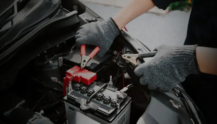 Mains de personne avec des gants gris connectant des câbles à une batterie de voiture. Un symptôme de batterie HS est souvent un démarrage difficile.