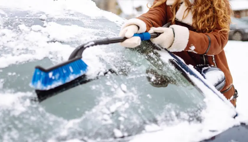 Une personne en manteau brun enlève la neige du pare-brise de sa voiture avec une brosse bleue. Faut il enlever la neige sur la voiture ? Oui, pour la sécurité !