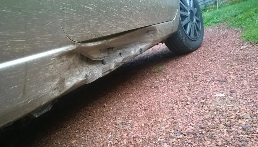 Vue rapprochée d'un bas de caisse enfoncé sur une voiture sale, montrant les dégâts et les fixations exposées.