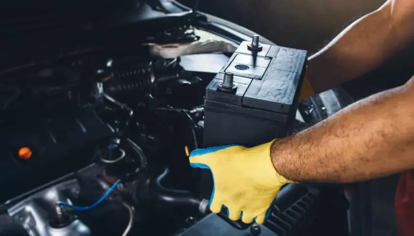 Mains pour gants jaunes tenant une batterie de voiture pour la débrancher. L'intérieur du compartiment moteur est visible.