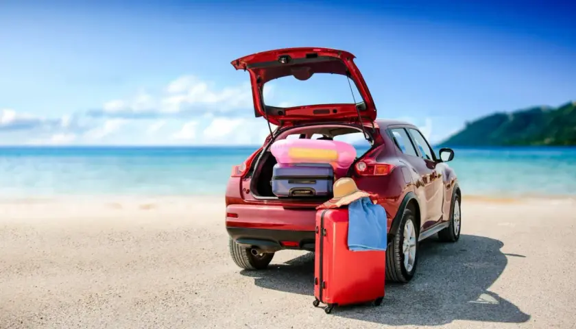 Voiture rouge prête pour le départ en vacances, coffre ouvert rempli de bagages, valise rouge et chapeau de paille sur le sable, mer bleue en arrière-plan.