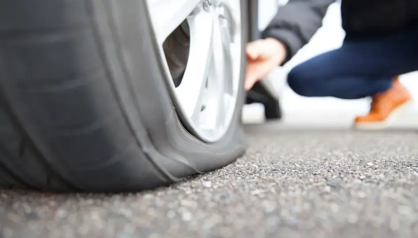 Gros plan sur un pneu crevé, une personne accroupie vérifie les dégâts. La route est asphaltée.